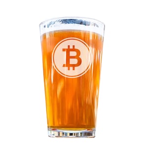 May include: A clear glass pint glass filled with a light amber colored beverage. The glass has a white Bitcoin logo printed on the side.