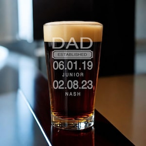 May include: A clear glass beer mug with a brown liquid inside. The mug is engraved with the text "DAD ESTABLISHED 06.01.19 JUNIOR 02.08.23 NASH".