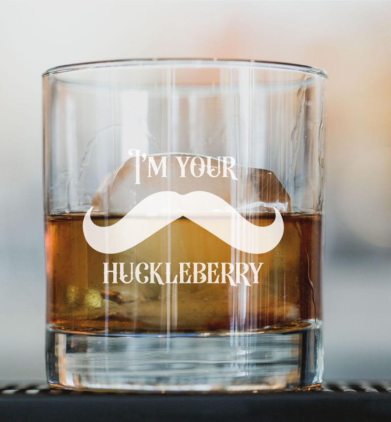 May include: A clear glass with a white mustache and the text "I'm your Huckleberry" printed on it. The glass is filled with a brown liquid and ice.