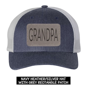 Könnte beinhalten: Marineblaue und weiße Trucker-Mütze mit einem grauen rechteckigen Aufnäher mit der Aufschrift "GRANDPA". Die Mütze hat einen Mesh-Rücken und einen gebogenen Schirm. Der Text "NAVY HEATHER/SILVER HAT WITH GREY RECTANGLE PATCH" steht unten.