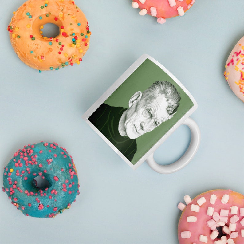 May include: White ceramic mug with a green background and a black and white illustration of a man's face. The mug has a handle on the right side.