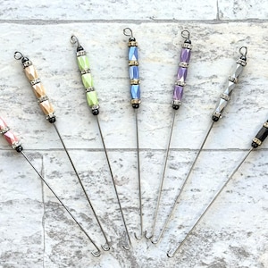 May include: A collection of seven decorative crochet hooks with colorful beaded handles. The handles feature various colors, including pink, beige, green, blue, purple, silver, and black, accented with silver and black details. Each hook has a silver metal shaft.