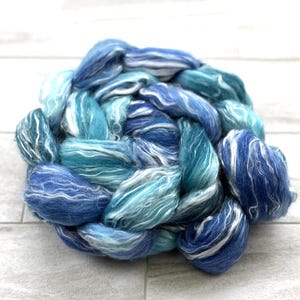 May include: A round bundle of hand-spun yarn in shades of blue and white. The yarn is arranged in a braided pattern, with varying tones of light and dark blue, and white highlights. The texture appears soft and fluffy, suitable for knitting or other fiber crafts.