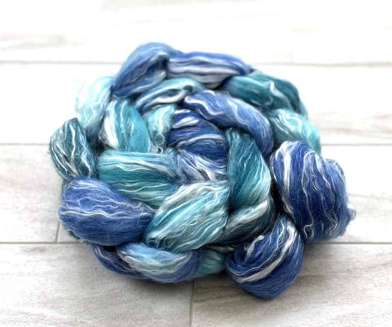 May include: A round bundle of hand-spun yarn in shades of blue and white. The yarn is arranged in a braided pattern, with varying tones of light and dark blue, and white highlights. The texture appears soft and fluffy, suitable for knitting or other fiber crafts.