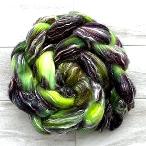 May include: A vibrant, hand-dyed fiber braid in shades of green, purple, black, and white. The fiber is tightly twisted, creating a textured, visually appealing bundle. Ideal for spinning, felting, or other fiber art projects.