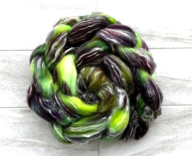 May include: A vibrant, hand-dyed fiber braid in shades of green, purple, black, and white. The fiber is tightly twisted, creating a textured, visually appealing bundle. Ideal for spinning, felting, or other fiber art projects.