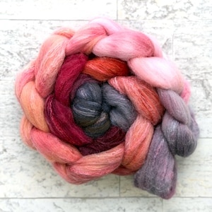 May include: A colorful braid of dyed wool roving in shades of pink, red, orange, and gray. The wool is arranged in a circular pattern, with the colors blending together. Ideal for fiber art projects.