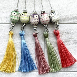 Pode incluir: Cinco alfinetes decorativos com contas de coruja em cerâmica verde, amarela, roxa, rosa e branca, cada um acentuado com pequenos strass transparentes e finalizado com uma longa borla de seda em amarelo, azul, malva, verde sálvia e vermelho coral.