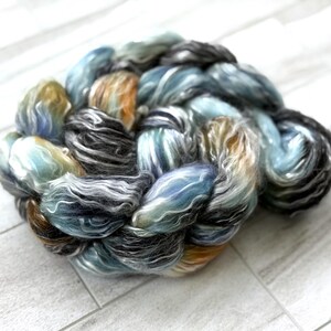 May include: A braided skein of hand-dyed yarn. The yarn displays a mix of colours, including blues, greys, and hints of orange and brown. The fibres look soft and fluffy, suitable for knitting or crochet projects.