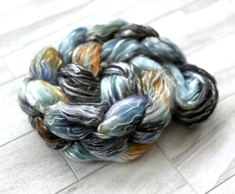May include: A braided skein of hand-dyed yarn. The yarn displays a mix of colours, including blues, greys, and hints of orange and brown. The fibres look soft and fluffy, suitable for knitting or crochet projects.
