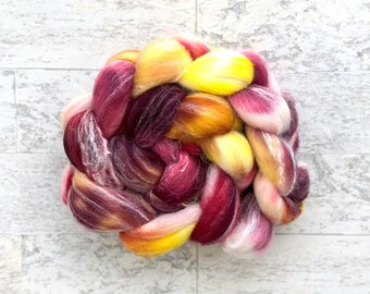 Può includere: Una colorata treccia di roving di lana tinto a mano nei toni del giallo, rosa e bordeaux. La fibra è disposta in una treccia spessa e strutturata, ideale per progetti di filatura o feltro. La lana ha un aspetto morbido e soffice.