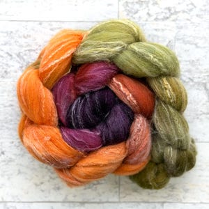 May include: A colorful braid of dyed wool roving in shades of orange, purple, red, and green. The fiber is arranged in a circular pattern, showcasing the blend of colors and textures. Ideal for fiber art projects.