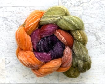 May include: A colorful braid of dyed wool roving in shades of orange, purple, red, and green. The fiber is arranged in a circular pattern, showcasing the blend of colors and textures. Ideal for fiber art projects.