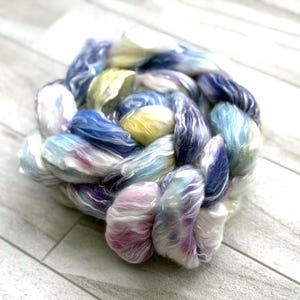 Op de afbeelding: Een streng handgeverfd garen in een vlecht. Het garen heeft een mix van kleuren, waaronder blauw, geel, roze en wit. De vezels zijn zacht en pluizig, ideaal voor brei- of haakprojecten.