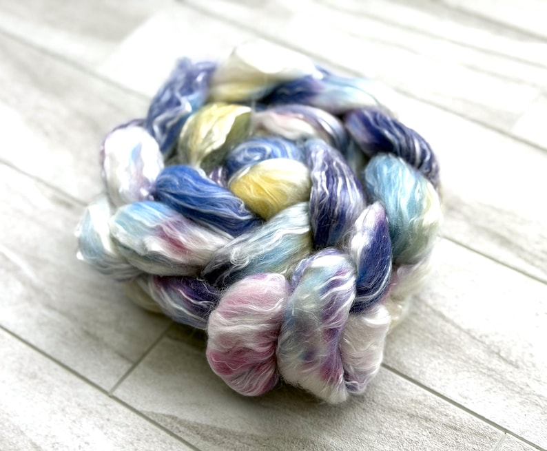 Op de afbeelding: Een streng handgeverfd garen in een vlecht. Het garen heeft een mix van kleuren, waaronder blauw, geel, roze en wit. De vezels zijn zacht en pluizig, ideaal voor brei- of haakprojecten.