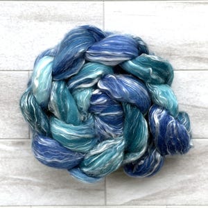 May include: A close-up of a braided bundle of blue and white fiber. The fiber is a mix of light and dark blues, with some white strands woven throughout. The texture appears soft and fluffy, ideal for crafting.