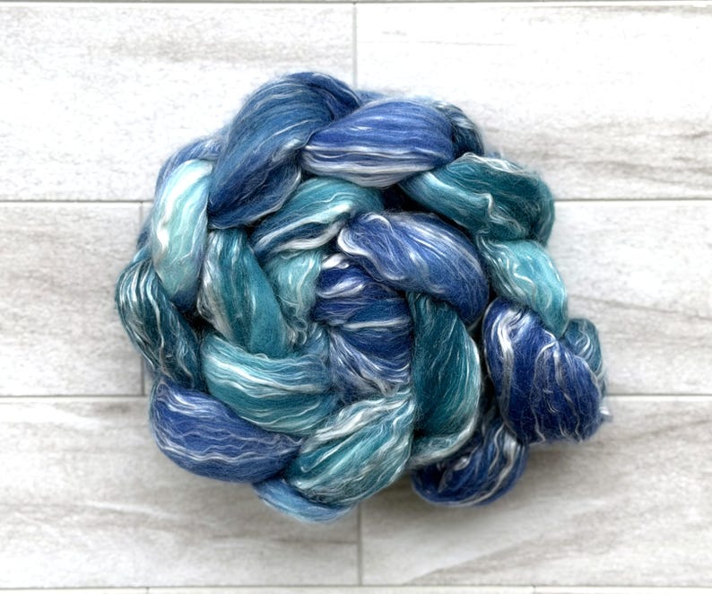 May include: A close-up of a braided bundle of blue and white fiber. The fiber is a mix of light and dark blues, with some white strands woven throughout. The texture appears soft and fluffy, ideal for crafting.