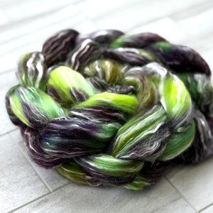 May include: A braided skein of hand-dyed yarn in shades of green, purple, and white. The yarn is arranged in a circular pattern, showcasing the blend of colors and textures. Ideal for knitting, crocheting, or other fiber arts projects.