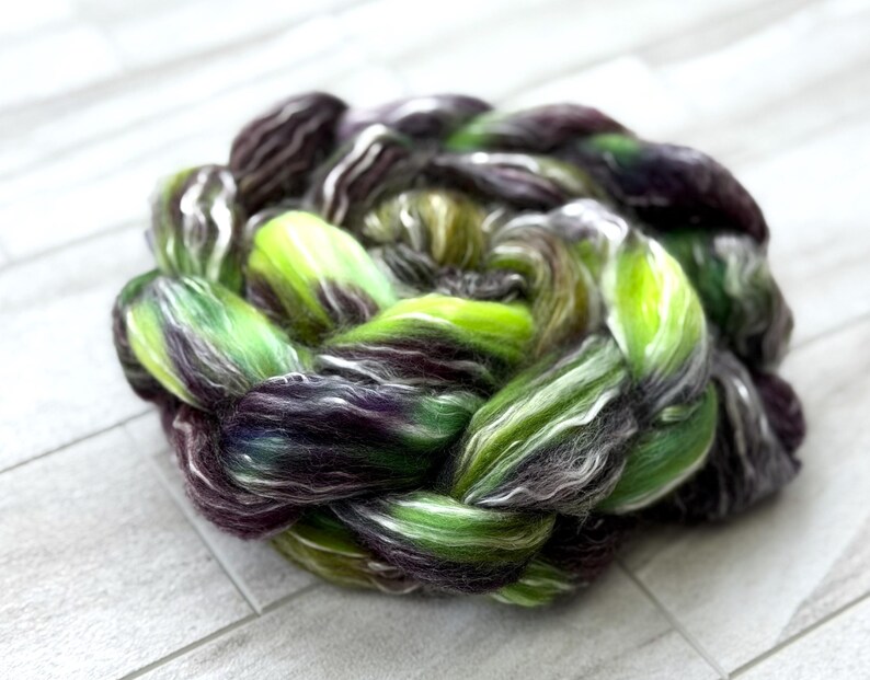 May include: A braided skein of hand-dyed yarn in shades of green, purple, and white. The yarn is arranged in a circular pattern, showcasing the blend of colors and textures. Ideal for knitting, crocheting, or other fiber arts projects.