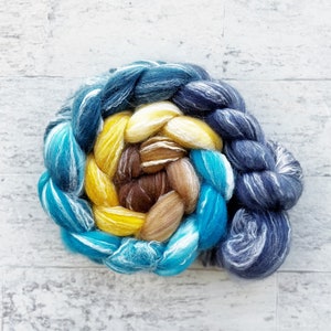 May include: A braided bundle of hand-dyed wool roving in shades of blue, yellow, brown, and white. The roving is arranged in a spiral pattern.