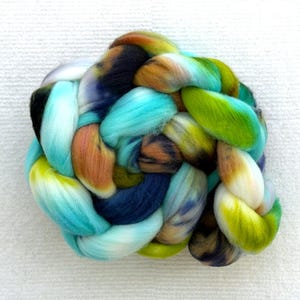 Puede incluir: Un paquete trenzado de mechas de lana coloridas. Las mechas presentan una mezcla de colores, incluyendo turquesa, verde, marrón y azul. La lana es suave y esponjosa, y la trenza está bien tejida. El fondo es blanco liso.
