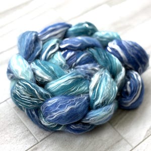 May include: A circular braid of hand-dyed wool roving in shades of blue and white. The roving is a mix of light and dark blues, with streaks of white throughout. The wool is soft and fluffy, perfect for spinning or felting projects.
