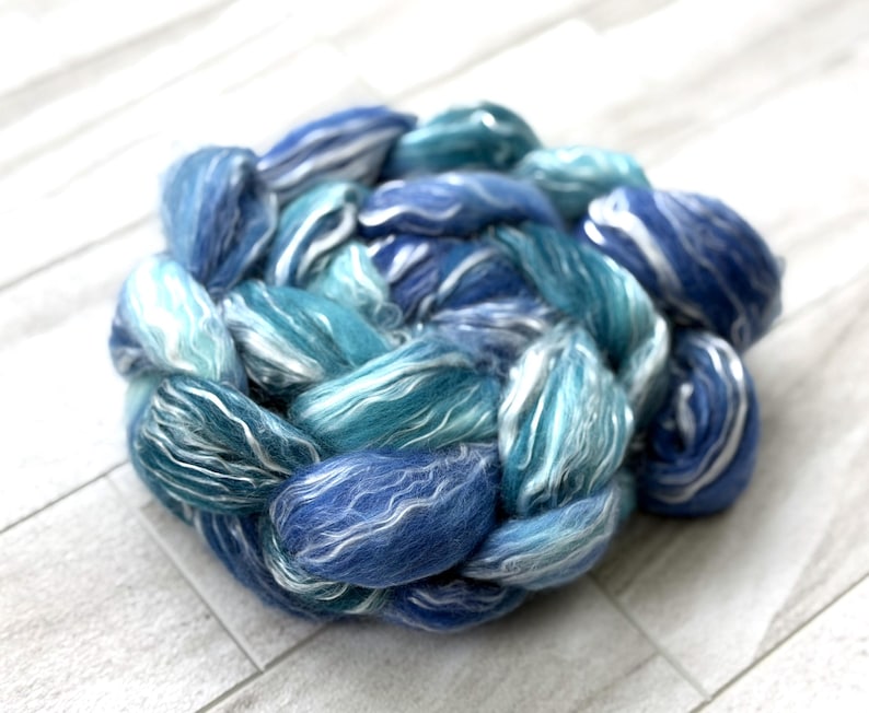 May include: A circular braid of hand-dyed wool roving in shades of blue and white. The roving is a mix of light and dark blues, with streaks of white throughout. The wool is soft and fluffy, perfect for spinning or felting projects.