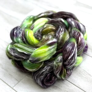 May include: A vibrant, hand-dyed fiber braid in shades of green, purple, and white. The fiber is tightly twisted, creating a textured, organic shape. The colors blend together, creating a unique and artistic look, perfect for spinning or other fiber arts.
