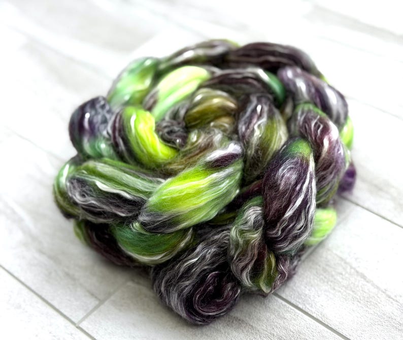 May include: A vibrant, hand-dyed fiber braid in shades of green, purple, and white. The fiber is tightly twisted, creating a textured, organic shape. The colors blend together, creating a unique and artistic look, perfect for spinning or other fiber arts.