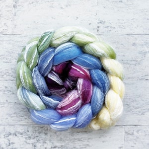 May include: A braided bundle of colorful yarn in shades of green, blue, purple, and yellow. The yarn is arranged in a spiral shape, with the ends of the braid tucked into the center.