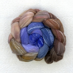 May include: A tightly braided bundle of colorful fiber in shades of brown, blue, and purple. The fiber is arranged in a circular pattern, with the colors blending together. The texture appears soft and fluffy, suggesting it is yarn or roving for crafts.