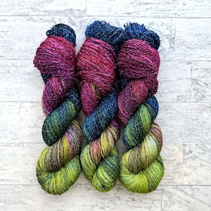 May include: Three skeins of yarn in shades of green, purple, and pink with silver glitter. The yarn is twisted into a decorative shape.