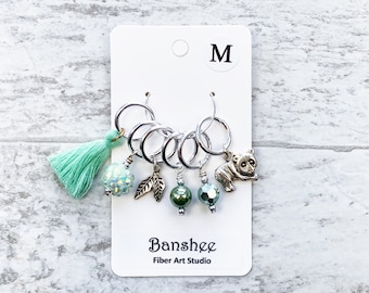 Puede incluir: Conjunto de seis marcadores de punto de plata con varios encantos, incluyendo un borla verde, una cuenta verde brillante, un encanto de hoja, una cuenta de perla verde, una cuenta verde brillante y un encanto de panda plateado. Los marcadores de punto se muestran en una tarjeta blanca con la letra "M" y el texto "Banshee Fiber Art Studio".
