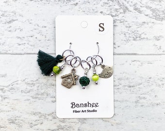 Puede incluir: Un juego de marcadores de punto con un borla verde, un amuleto de casita de pájaros, una cuenta verde y plateada, una cuenta amarilla y un amuleto de pájaro. Los marcadores de punto están en una tarjeta blanca con el texto "Banshee Fiber Art Studio" y "S".