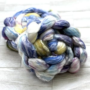 Op de afbeelding: Een gevlochten streng kleurrijk garen in tinten blauw, paars, geel en wit. Het garen is strak geweven, waardoor een textuurrijk, tastbaar uiterlijk ontstaat. De kleuren vloeien in elkaar over, waardoor een zacht aquareleffect ontstaat.