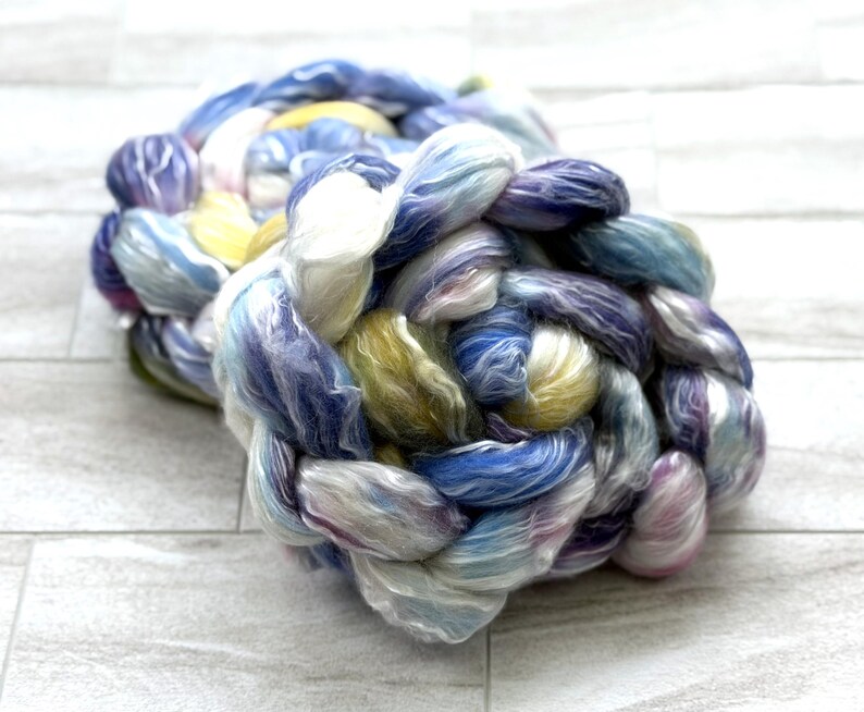 Op de afbeelding: Een gevlochten streng kleurrijk garen in tinten blauw, paars, geel en wit. Het garen is strak geweven, waardoor een textuurrijk, tastbaar uiterlijk ontstaat. De kleuren vloeien in elkaar over, waardoor een zacht aquareleffect ontstaat.