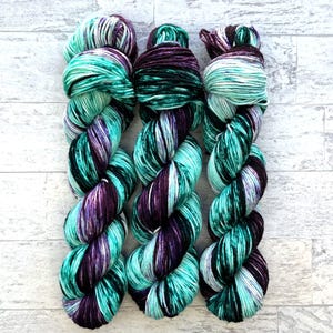 Op de afbeelding: Drie bollen garen in een teal, paarse en zwarte kleurstelling. Het garen is gedraaid en klaar om te gebruiken voor breien of haken.