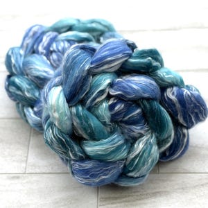 May include: A braided bundle of fiber in shades of blue and teal, with white highlights. The fiber appears soft and fluffy, with a textured appearance. The bundle is arranged in a circular shape, suggesting it is for crafting or textile projects.