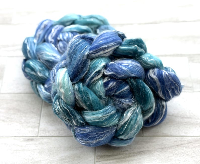 May include: A braided bundle of fiber in shades of blue and teal, with white highlights. The fiber appears soft and fluffy, with a textured appearance. The bundle is arranged in a circular shape, suggesting it is for crafting or textile projects.