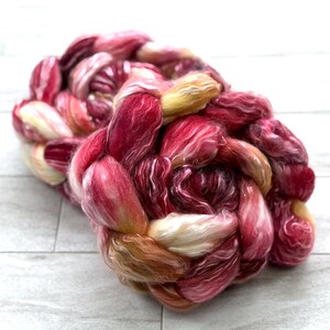 May include: A braided bundle of colorful yarn in shades of red, pink, and yellow. The yarn appears soft and fluffy, with a mix of light and dark tones. The fibers are tightly woven together, creating a textured appearance. This yarn is suitable for knitting or other fiber crafts.