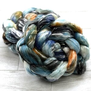 May include: A close-up of a braided skein of colourful yarn. The yarn features a blend of blues, greens, oranges, and greys. The fibres are tightly twisted, creating a textured appearance. The yarn is likely used for knitting or other fibre crafts.
