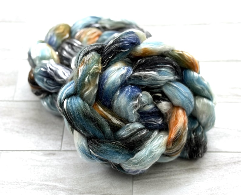 May include: A close-up of a braided skein of colourful yarn. The yarn features a blend of blues, greens, oranges, and greys. The fibres are tightly twisted, creating a textured appearance. The yarn is likely used for knitting or other fibre crafts.