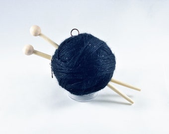 Puede incluir: Una madeja de lana azul oscuro con motas plateadas, sobre una base de plástico transparente. Dos agujas de tejer de madera están insertadas en la lana.