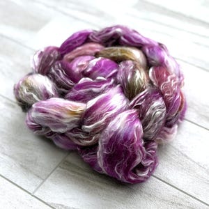 Puede incluir: Una trenza de fibra te&ntilde;ida a mano en tonos vibrantes de morado, rosa y blanco. La fibra est&aacute; dispuesta en forma circular, mostrando la mezcla de colores y texturas. Ideal para hilar, tejer u otros proyectos de arte textil.
