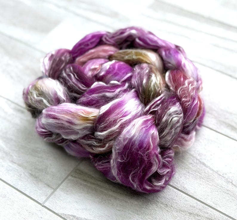 Puede incluir: Una trenza de fibra te&ntilde;ida a mano en tonos vibrantes de morado, rosa y blanco. La fibra est&aacute; dispuesta en forma circular, mostrando la mezcla de colores y texturas. Ideal para hilar, tejer u otros proyectos de arte textil.
