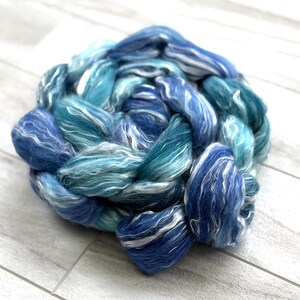 May include: A close-up of a skein of hand-dyed fiber in shades of blue and teal with white highlights. The fiber is arranged in a circular shape, with the strands of wool twisted together. The color palette evokes the ocean.