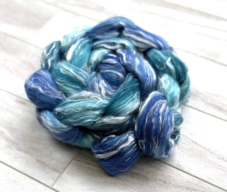May include: A close-up of a skein of hand-dyed fiber in shades of blue and teal with white highlights. The fiber is arranged in a circular shape, with the strands of wool twisted together. The color palette evokes the ocean.