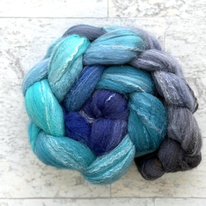 May include: A braided skein of hand-dyed yarn in shades of blue and teal. The yarn is arranged in a circular pattern, with the colors transitioning from light teal to deep indigo and gray. The texture of the yarn appears soft and fluffy.