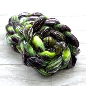 May include: Braided yarn in shades of green, purple, and white. The yarn is tightly woven, creating a textured, rope-like appearance. The colors blend together, creating a unique and visually appealing fiber product.