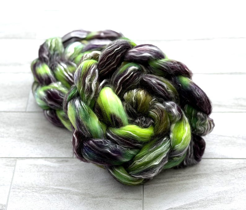 May include: Braided yarn in shades of green, purple, and white. The yarn is tightly woven, creating a textured, rope-like appearance. The colors blend together, creating a unique and visually appealing fiber product.